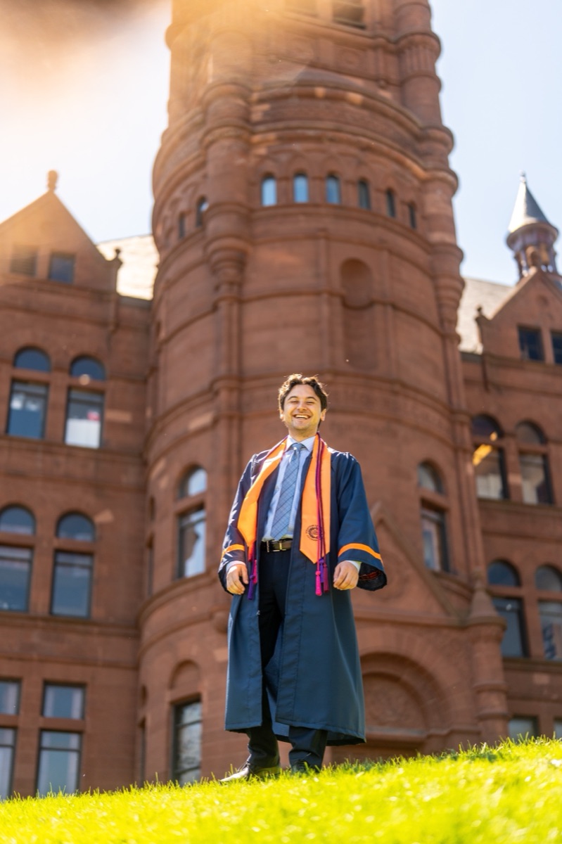 Graduate in a navy gown and orange stole laughing in front of Crouse College at Syracuse University