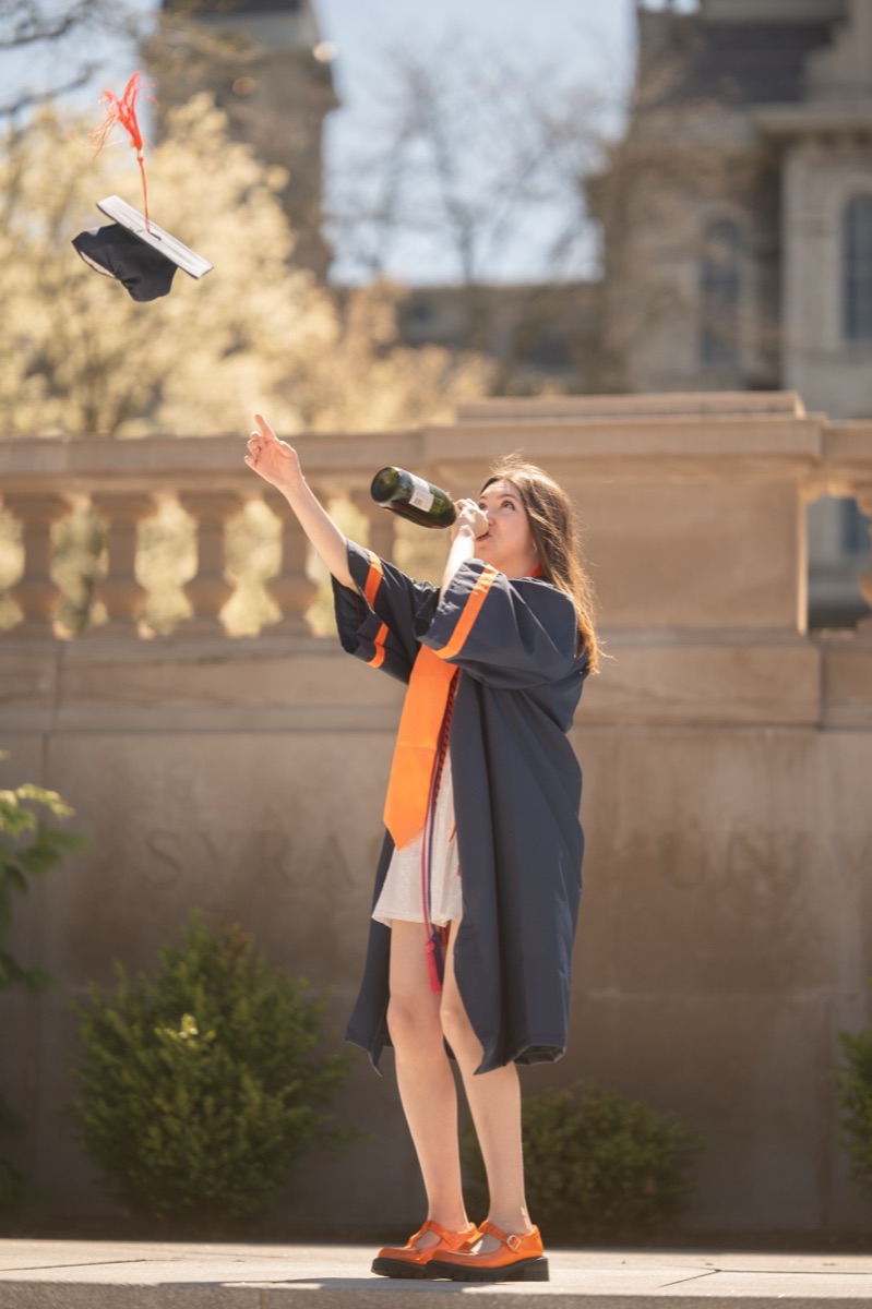 Graduate sipping champagne while flinging her cap in front of a carved Syracuse University stone wall