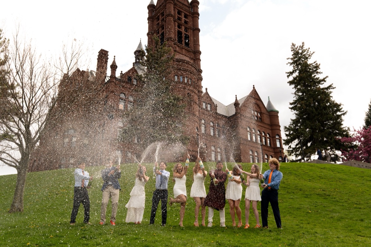 Friends in graduation whites spraying champagne on the lawn below Crouse College