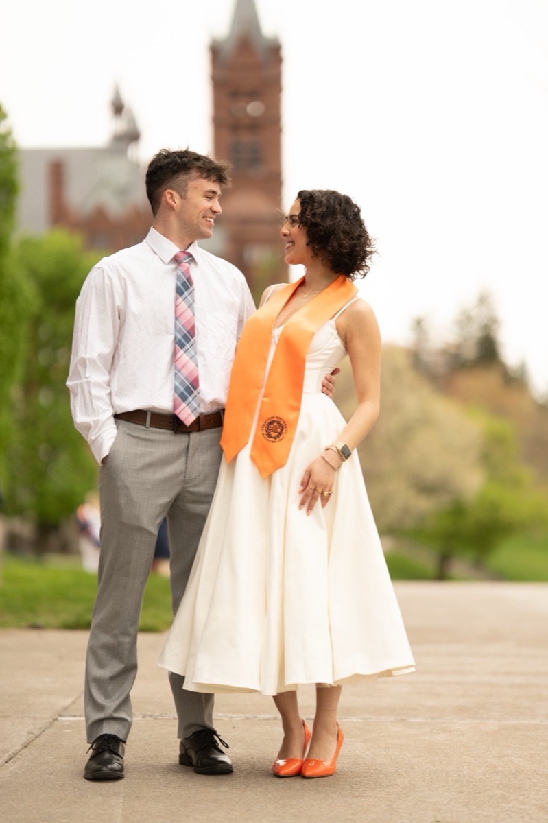 Couple sharing a smile on the campus promenade with the Crouse College tower behind them