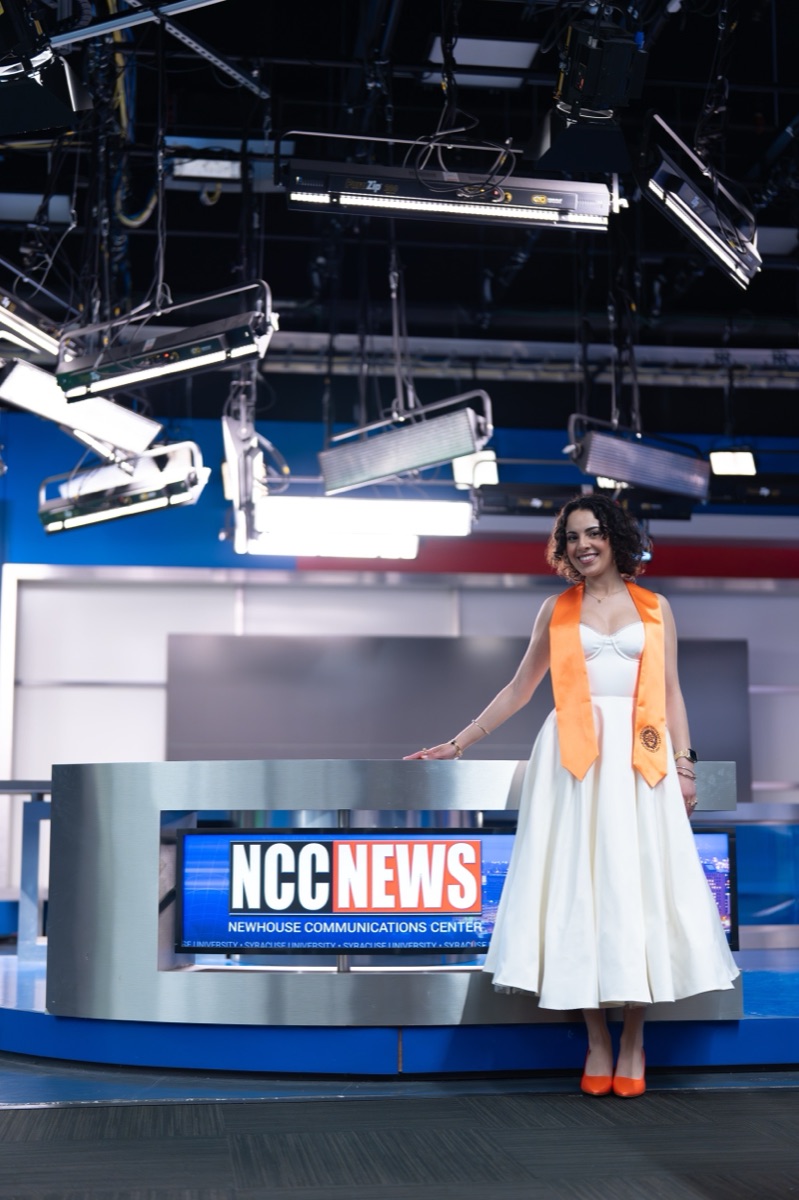 Newhouse graduate posing in the NCC News studio beneath the broadcast lighting grid