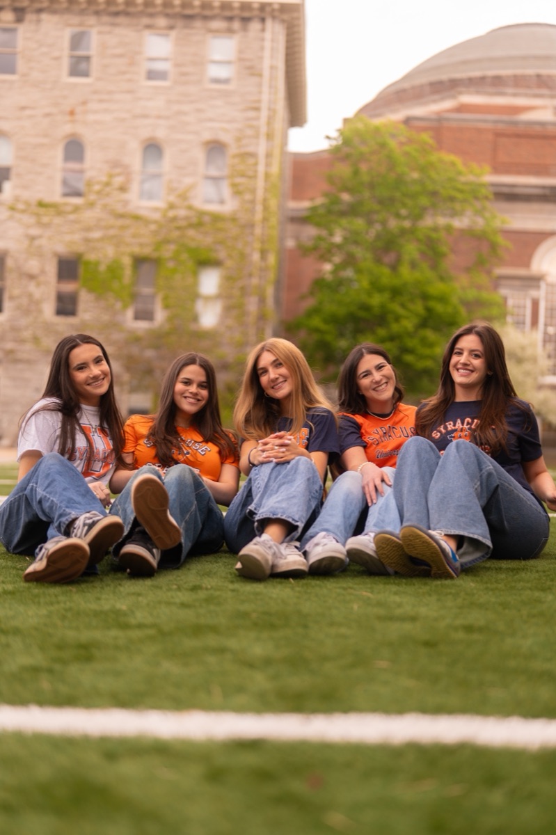 Five friends in Syracuse shirts relaxing together on the campus turf field