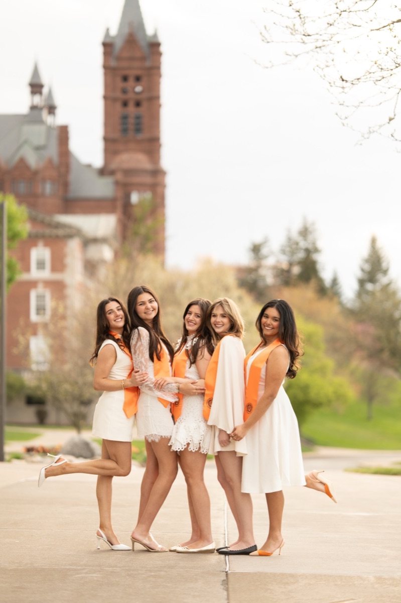 Five graduates in white dresses and orange stoles posing together on the walkway below Crouse College