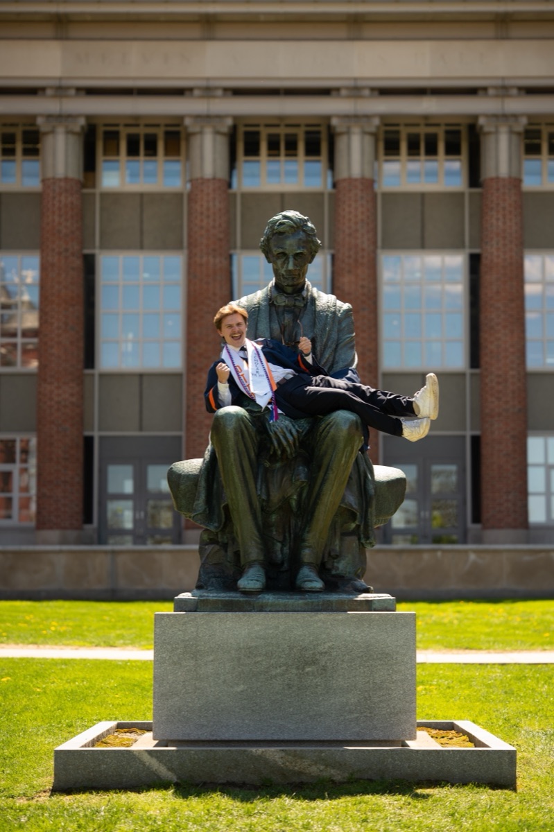 Graduate posing playfully in the lap of the Lincoln statue on the Syracuse campus