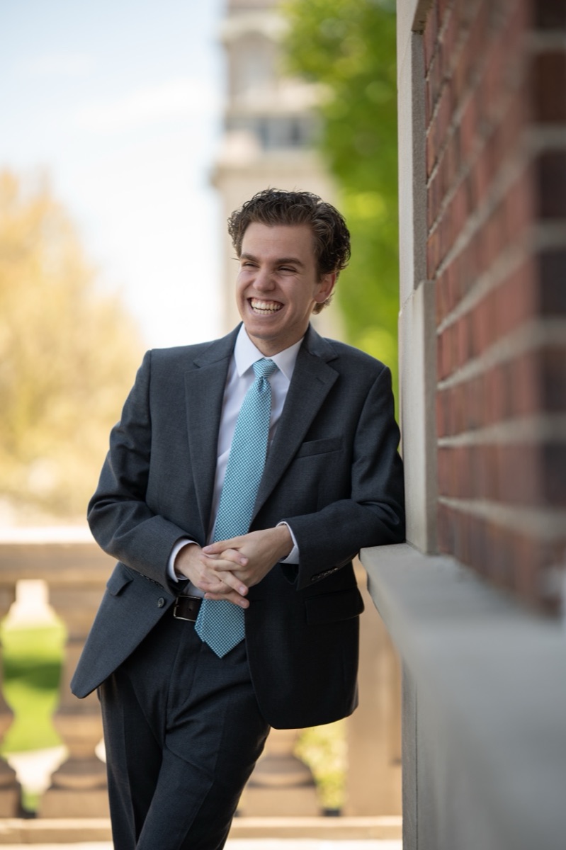 Graduate in a charcoal suit and teal tie laughing beside a brick archway