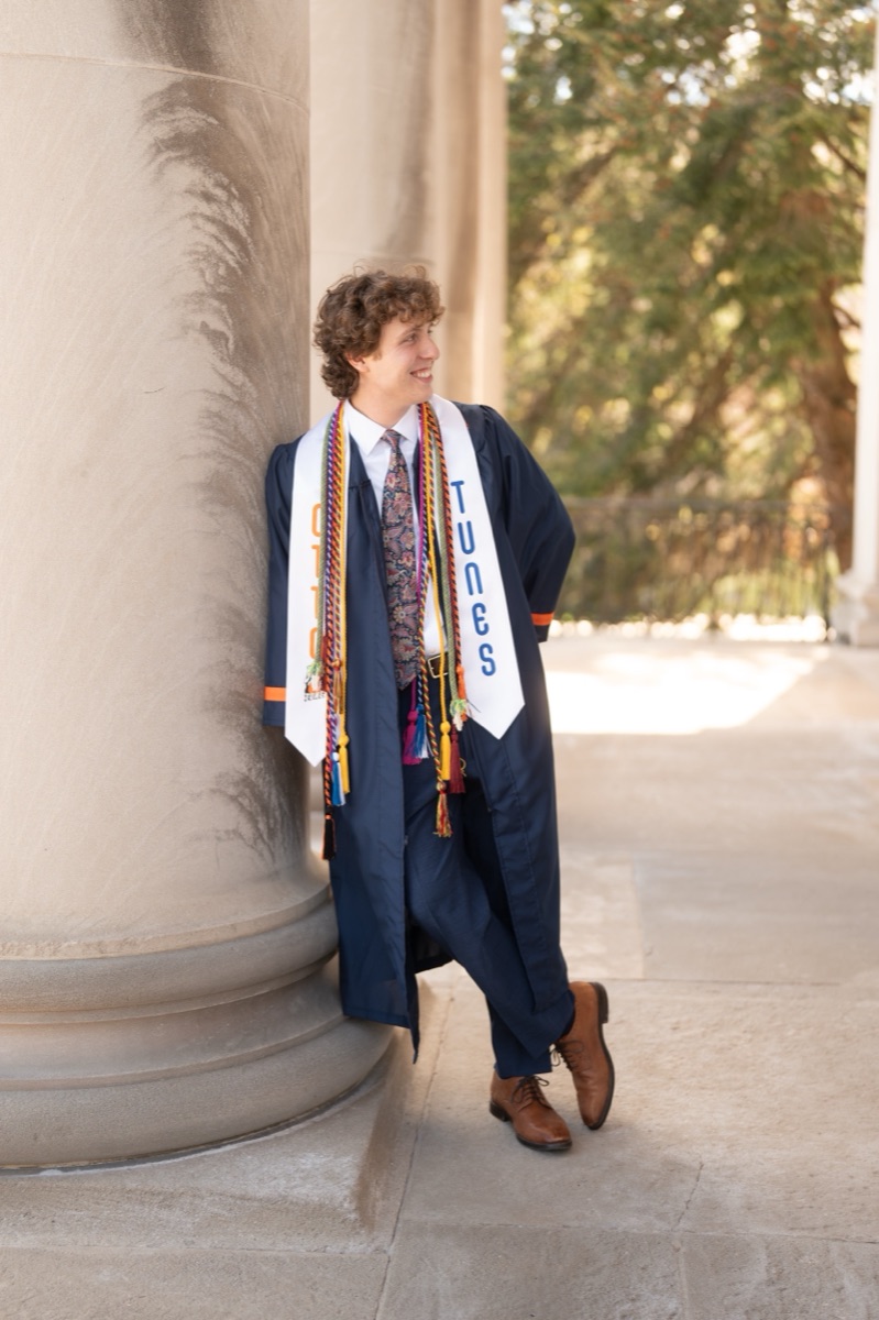 Otto Tunes singer in his gown, stole, and honor cords leaning against a limestone column