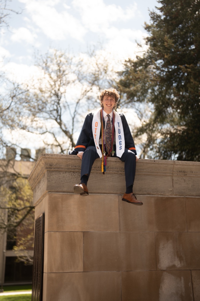 Graduate with honor cords and an Otto Tunes stole sitting atop a stone monument