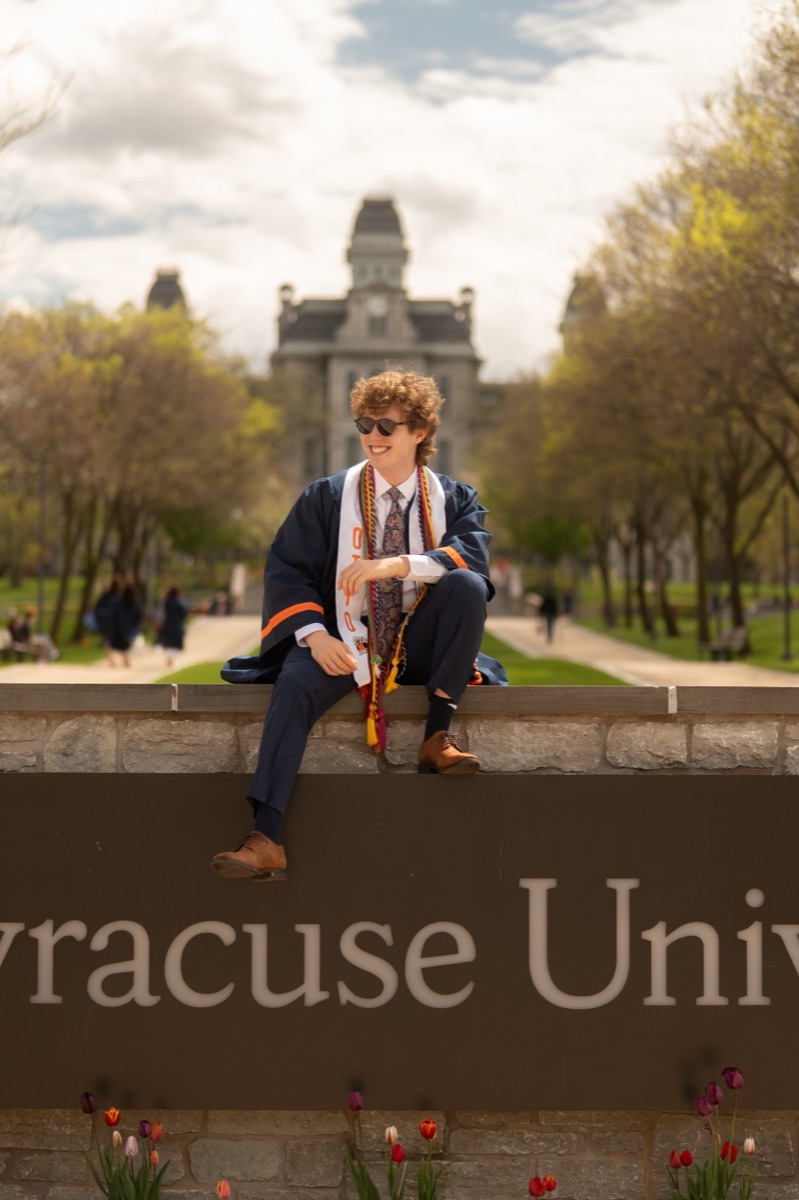 Graduate in sunglasses sitting on the Syracuse University entrance sign among spring tulips